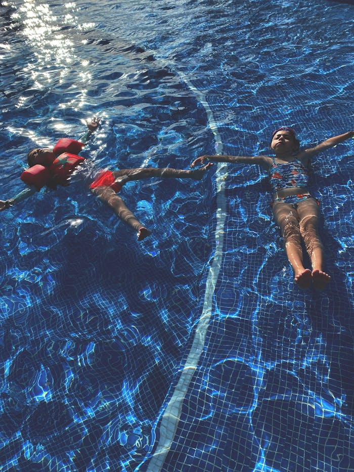pexels-photo-11114684 Two children relaxing and floating in a swimming pool with water wings under bright sunlight.